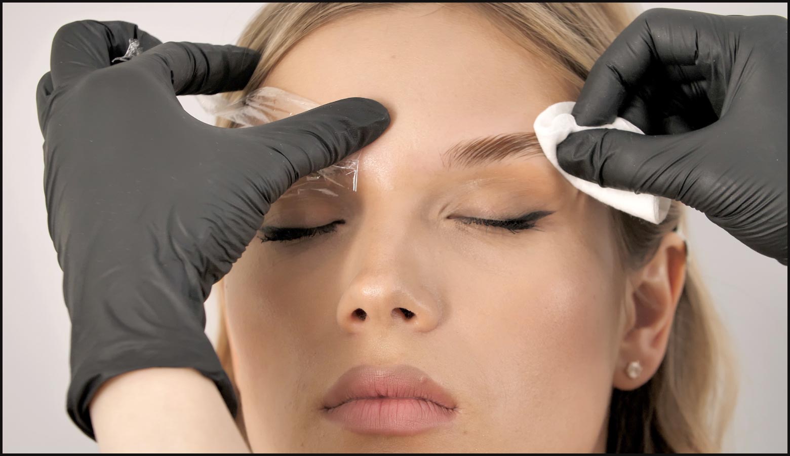 close up on client’s face with eyes closed, brow artist’s gloved hands are cleaning eyebrows 